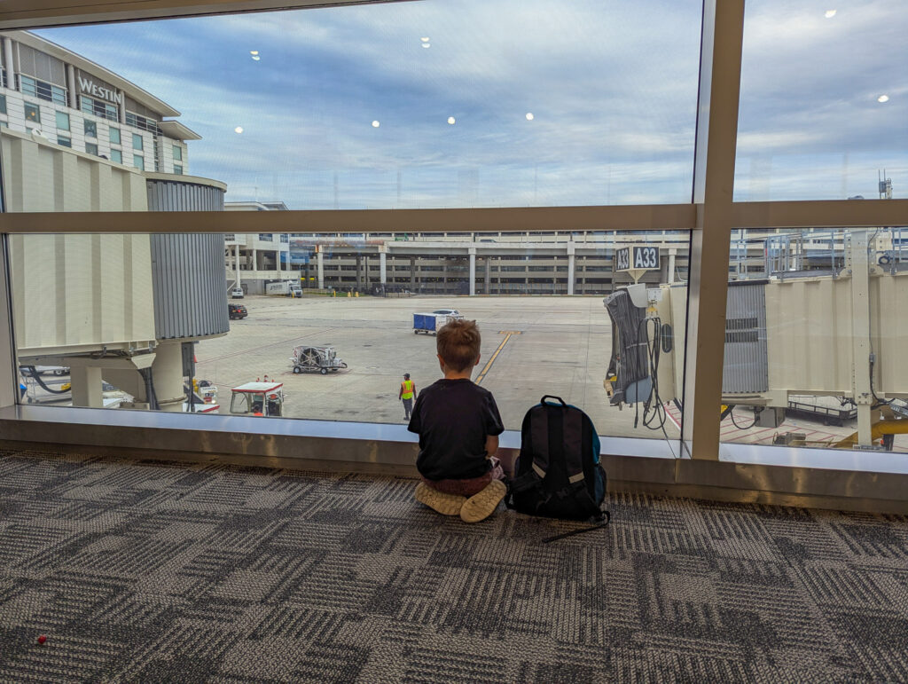 Child sits on airport terminal floor watching tarmac activity through large windows during overcast daytime conditions
