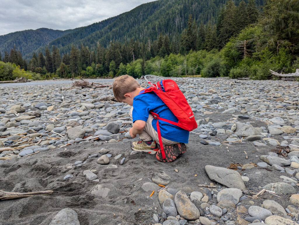 A young hiker with red backpack crouches on rocky Hoh river riverbank examining stones in forested Pacific Northwest valley during overcast day