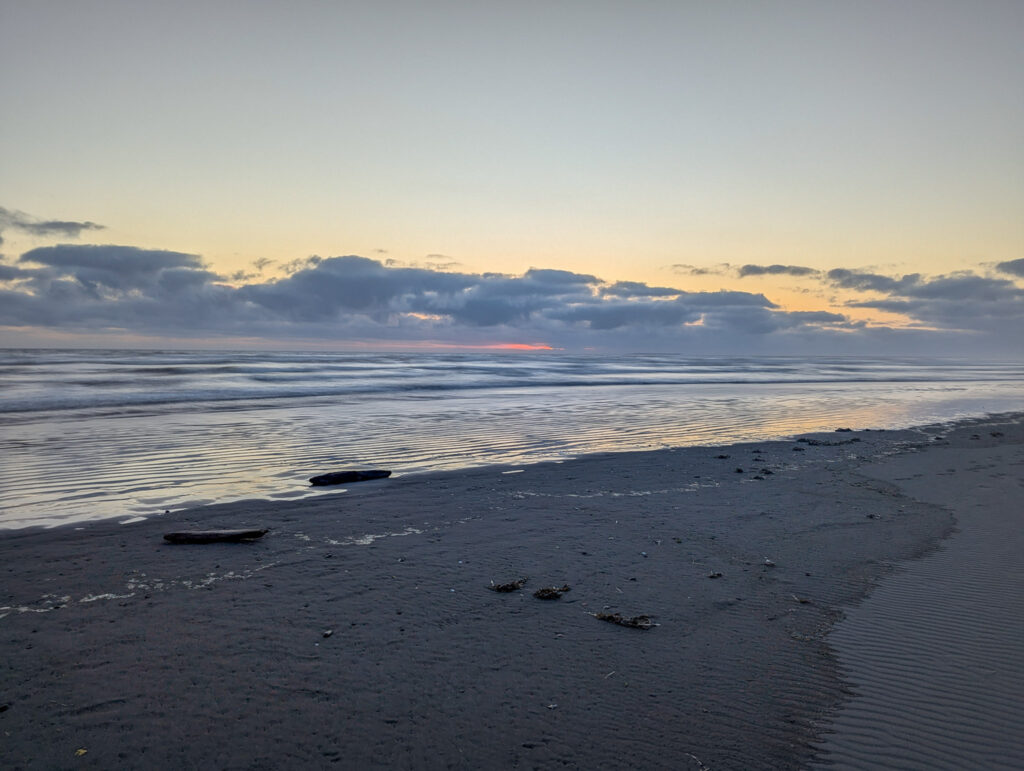 Twilight beach scene with dark sand, gentle waves, driftwood, and clouds illuminated by sunset colors on the horizon at Kalaloch Beach
