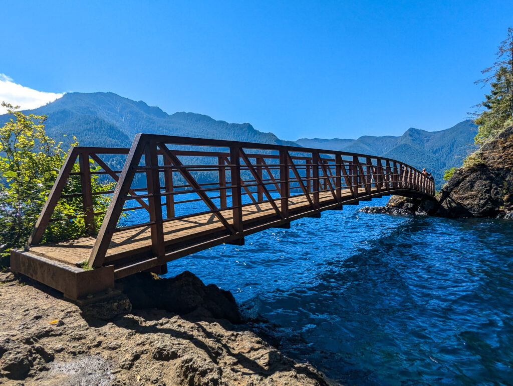 Steel footbridge crosses over Devil's Punchbowl in Lake Crescent through forested mountain valley under clear afternoon sky in Olympic National Park
