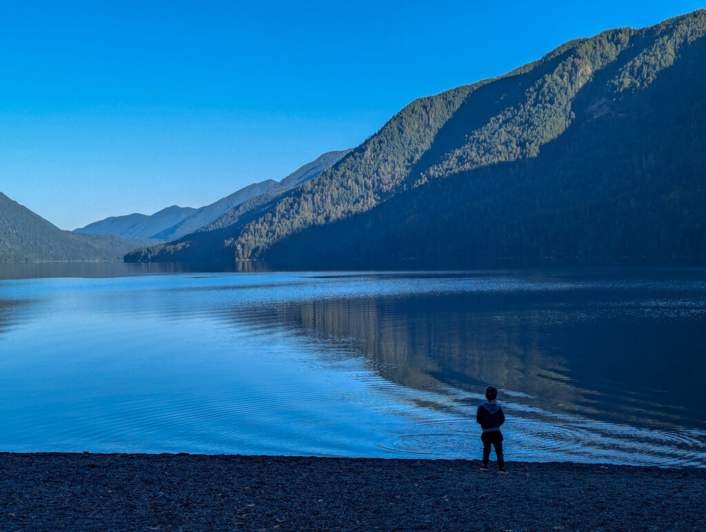 child standing at edge of mountain lake with forested slopes reflected in calm blue water during golden hour