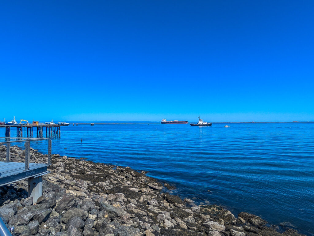 Rocky coastline with wooden pier and cargo ships anchored in calm blue harbor waters under clear daytime sky in Port Townsend, Washington