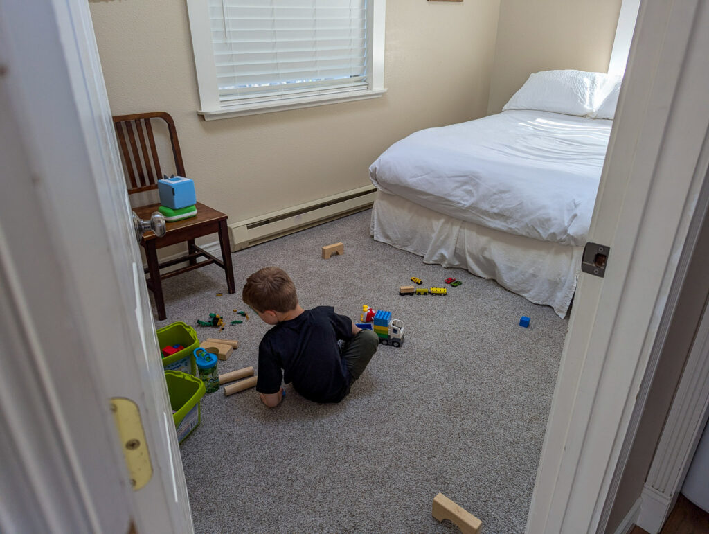 Child playing with toys on carpet in small bedroom with twin bed, wooden chair, and window with blinds