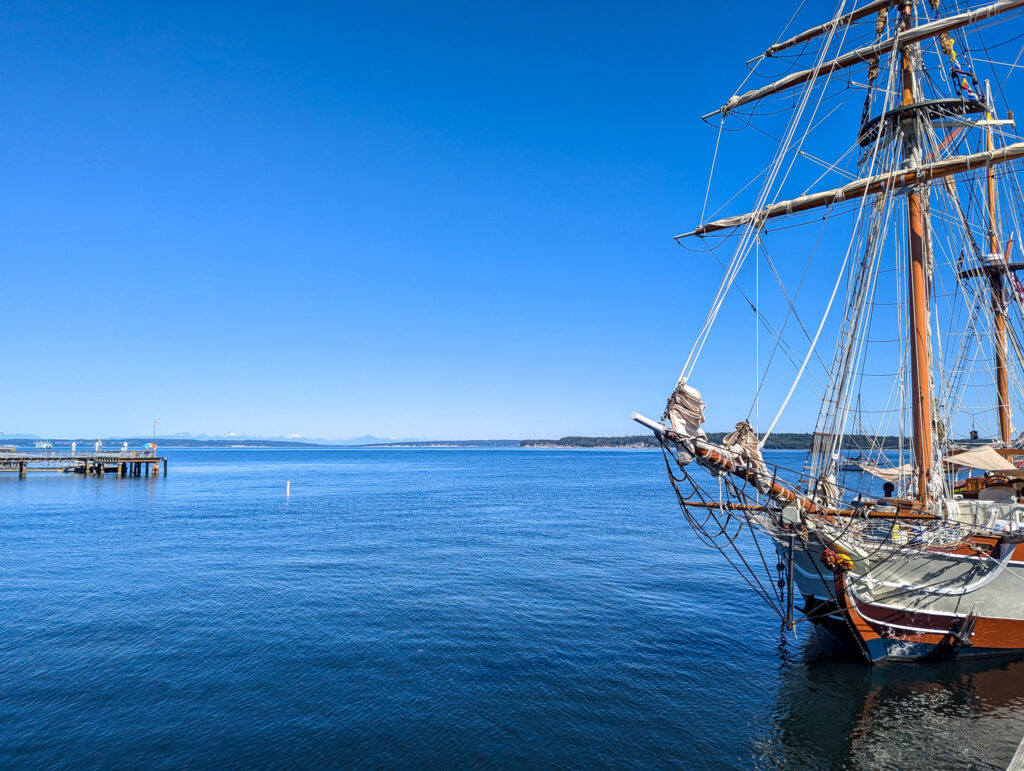 Historic tall ship with furled sails docked in calm blue harbor waters under clear daytime sky with distant pier visible in Port Townsend, Washington