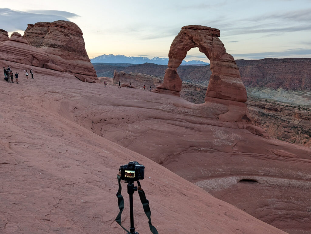 A camera on a tripod faces Delicate Arch at golden hour, with snow-capped La Sal Mountains framed through the opening.