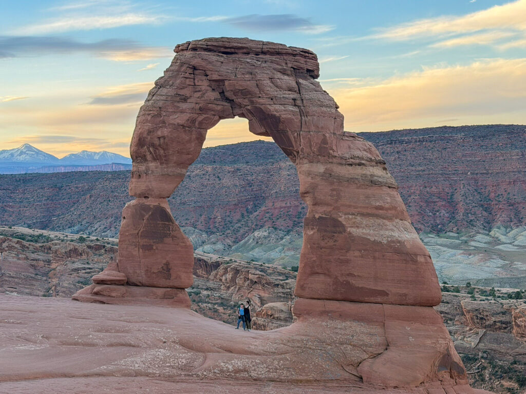 Two visitors stand beneath Delicate Arch at golden hour, with snow-capped La Sal Mountains visible in the distance.