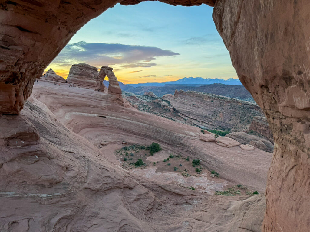 Delicate Arch seen through a sandstone alcove at golden hour, with the snow-capped La Sal Mountains glowing on the horizon.