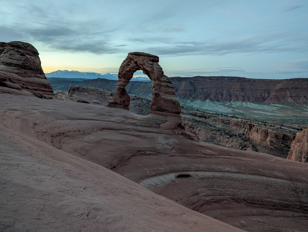 Delicate Arch rises above a sweeping slickrock bowl at dawn, with the Colorado River canyon and La Sal Mountains framed in the opening.