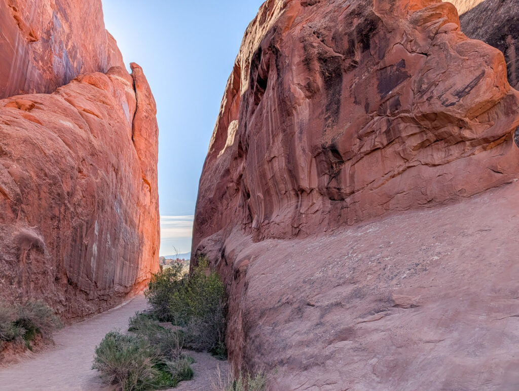 Narrow slot between towering red Entrada sandstone fins in Arches National Park's Devils Garden, with blue sky above.