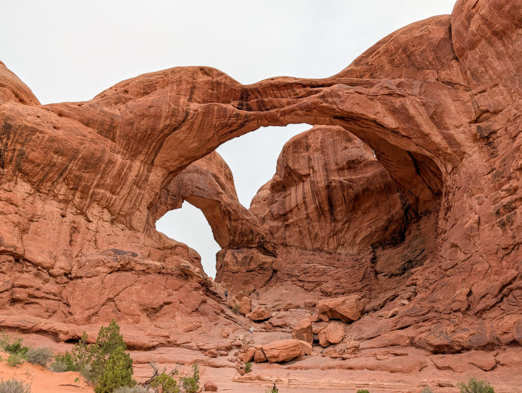 Front-on view of Double Arch in Arches National Park showing both sandstone spans under overcast skies, with tiny visitors visible at the base.