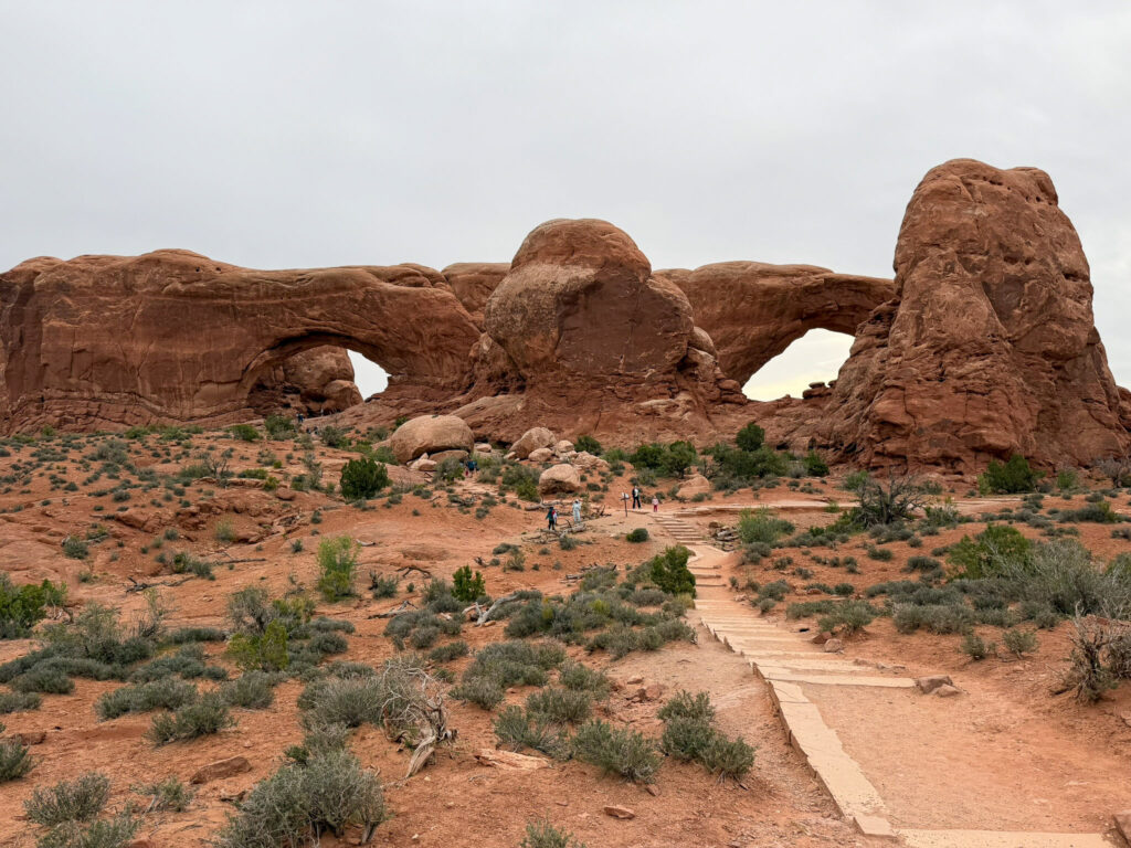 Paved Windows Trail leading toward North Window and South Window arches framing a red rock fin in Arches National Park.