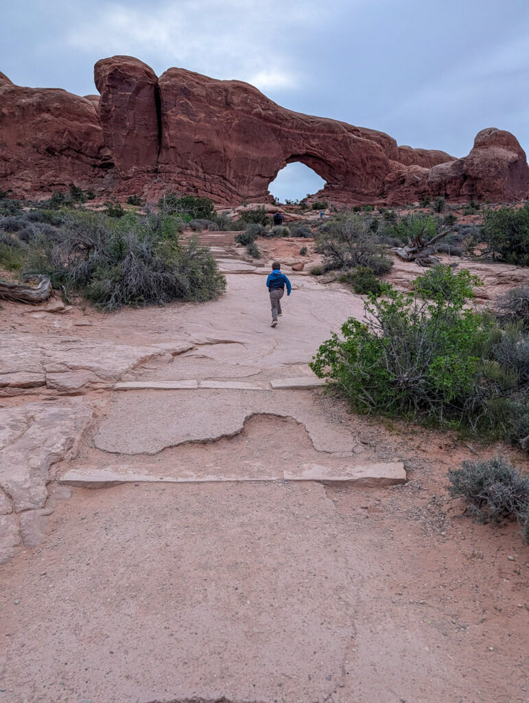 A child in a blue hoodie runs along the sandstone trail toward North Window Arch under an overcast sky in Arches National Park.