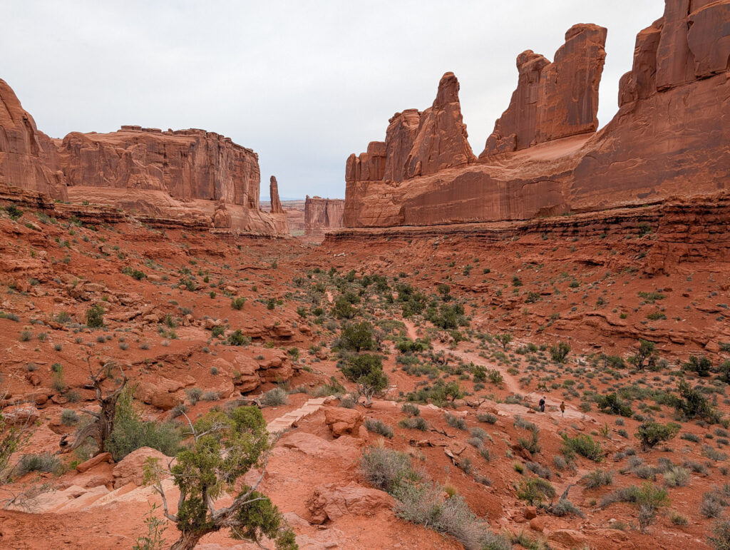 Two hikers walk the Park Avenue Trail corridor in Arches National Park, flanked by towering red sandstone fins under overcast skies.
