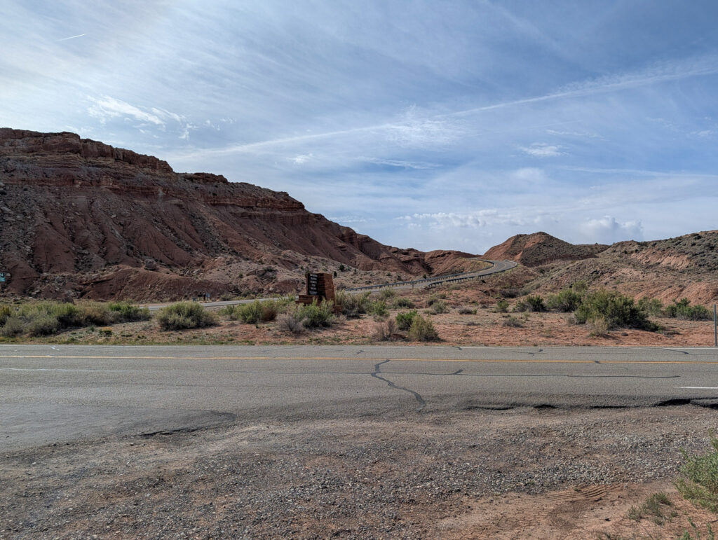 Capitol Reef National Park entrance sign stands along a two-lane highway, with towering red sandstone cliffs rising behind it.
