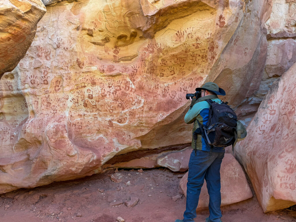 A hiker photographs the Fremont Culture Handprint Pictograph Panel, a sandstone wall densely covered in dark red hand stencils near Capitol Reef.