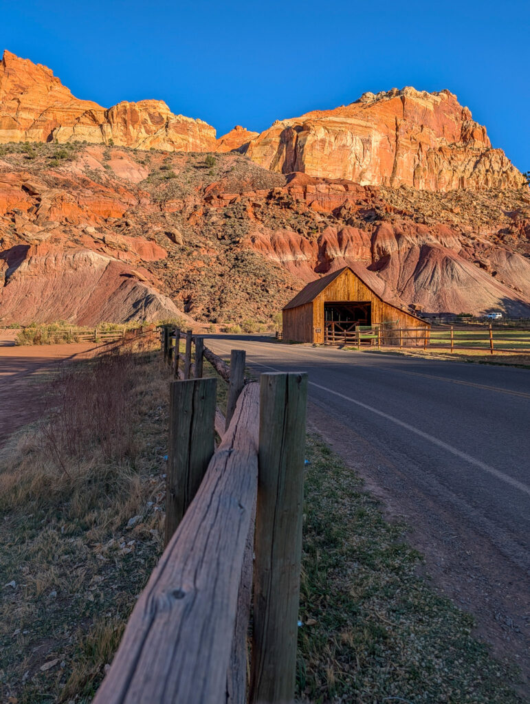 Historic Gifford Homestead Barn stands beside a split-rail fence with towering red sandstone cliffs rising behind it in Capitol Reef.