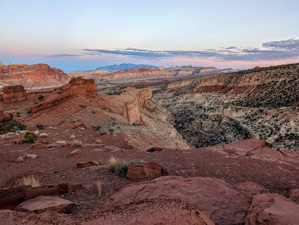 Panoramic dusk view from Sunset Point Overlook in Capitol Reef, with layered red-rock canyons, the Waterpocket Fold, and the distant Henry Mountains under a pastel twilight sky.
