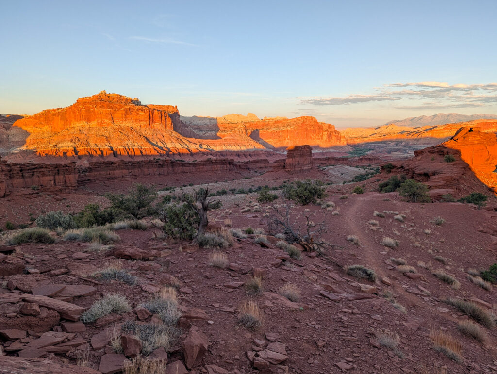 Wide evening view from Sunset Point in Capitol Reef, with orange-red Waterpocket Fold cliffs glowing under low light above a winding desert trail.