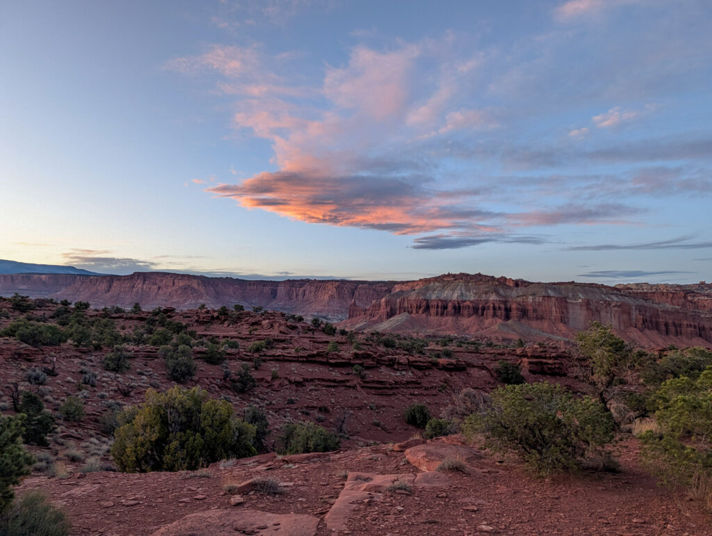 Panoramic dusk view from Capitol Reef's Waterpocket Fold Overlook, with layered red sandstone cliffs beneath pink and orange alto-cumulus clouds.