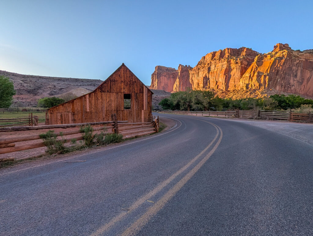 Historic Gifford Homestead barn along Capitol Reef's Scenic Drive, with golden sandstone cliffs rising behind split-rail fences and a curving road.