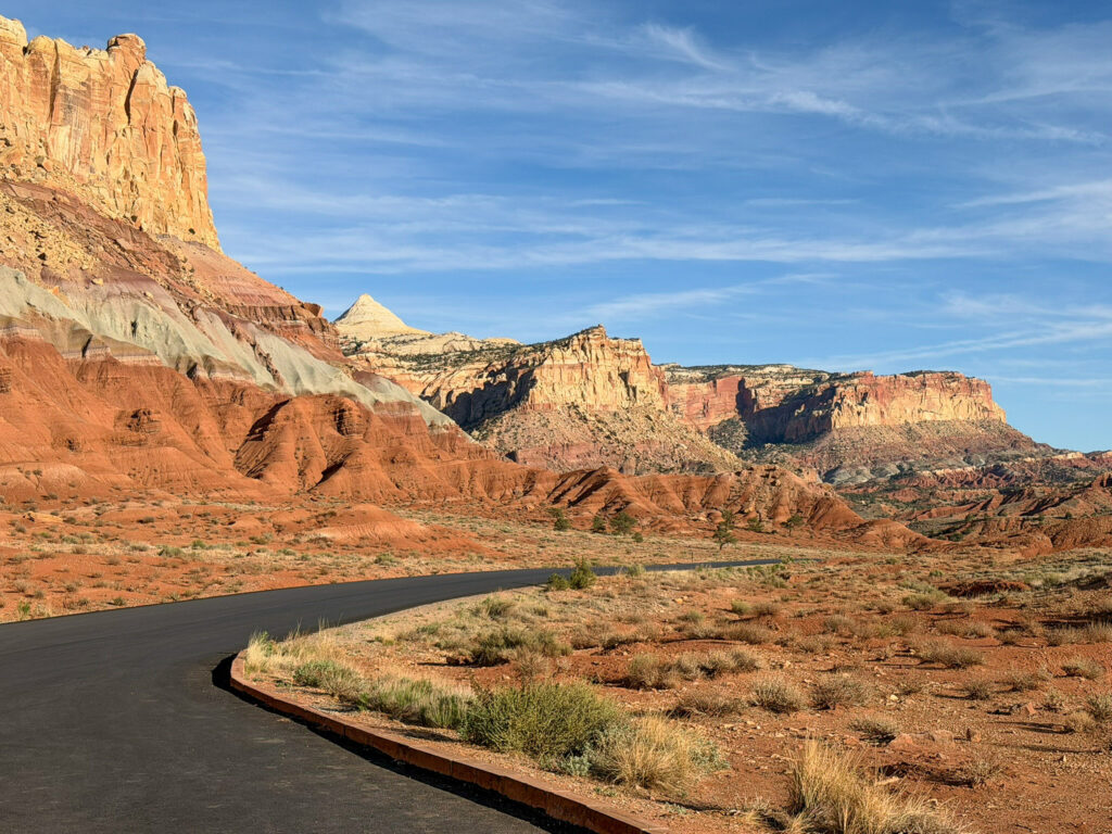 Capitol Reef Scenic Drive curves through red desert scrubland beneath layered sandstone cliffs glowing in warm late-day light.