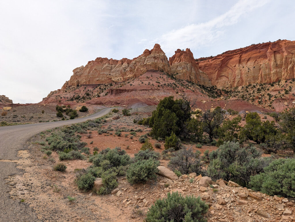 Two-lane road winds through Capitol Reef's Waterpocket Fold beneath layered red and cream sandstone cliffs under overcast skies.