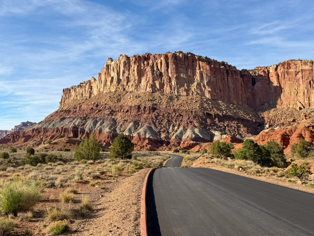 Two-lane road winds toward the towering layered sandstone cliffs of Capitol Reef Scenic Drive at blue hour, flanked by desert scrub and junipers.