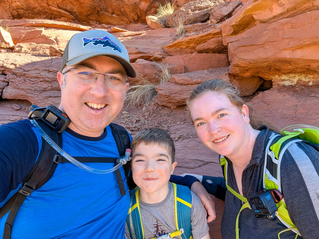 Family of three hikers pose for selfie against red sandstone rock formations in desert canyon during bright daylight