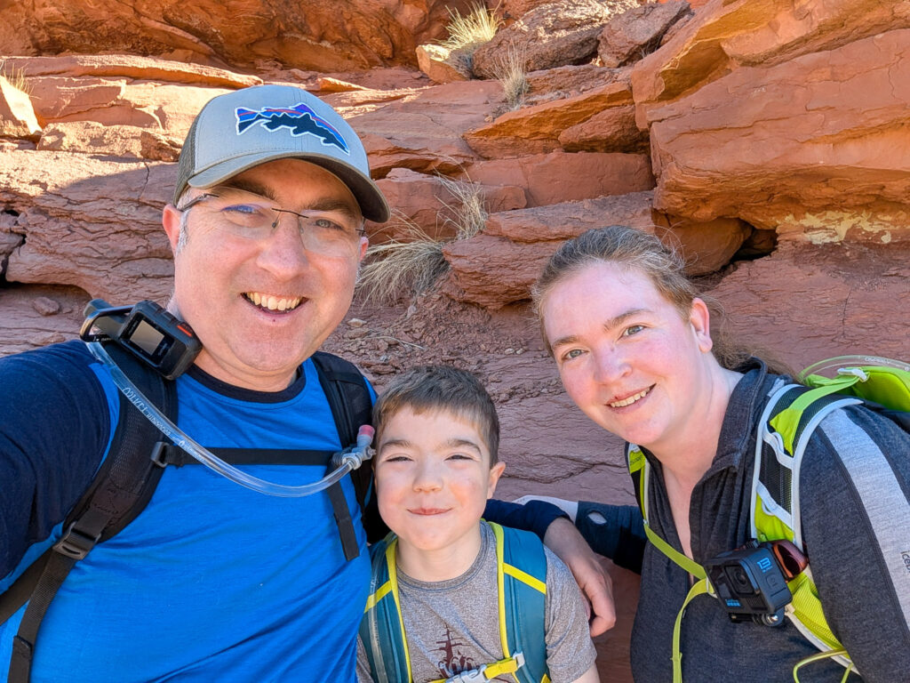 A family of three takes a selfie in a narrow red sandstone canyon alcove along a Capitol Reef National Park trail.