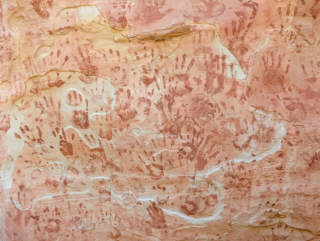 Dense panel of red ochre hand prints and finger dots in Barrier Canyon Style pictographs on a pale sandstone wall at Capitol Reef National Park.