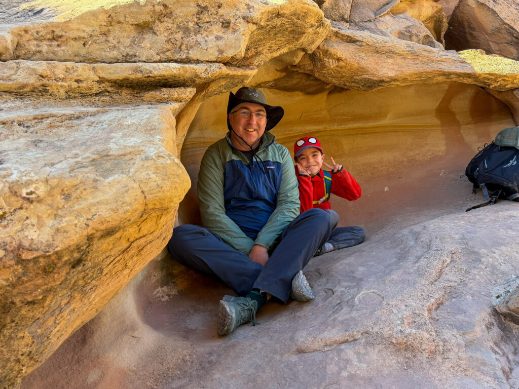 A father and child rest inside a smooth Navajo sandstone alcove along a Capitol Reef National Park trail, the child flashing a peace sign.