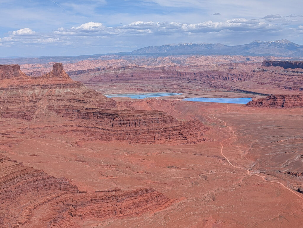 Wide canyon view from Dead Horse Point Overlook with the Colorado River below and turquoise Intrepid Potash evaporation ponds in the distance, framed by red rock walls and the snow-capped La Sal Mountains.