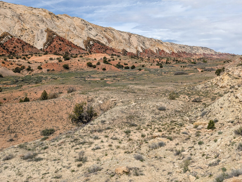 Wide valley view toward the Cockscomb monocline in Grand Staircase-Escalante, its red Carmel Formation and white Navajo Sandstone layers stretching across the horizon above pale caliche flats.