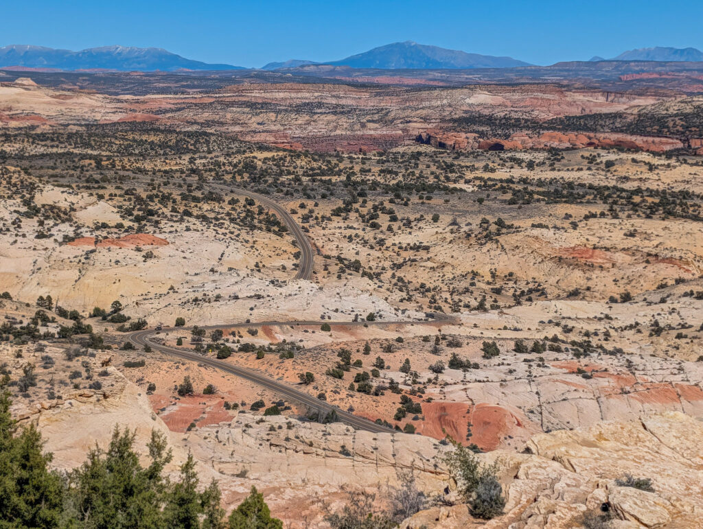 Highway 12 winds through dramatic S-curves of cream and rust slickrock at the Head of the Rocks Overlook, with snow-capped Henry Mountains on the horizon.