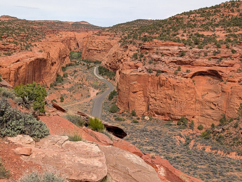 Cottonwood Canyon Road winds through a narrow red-rock canyon, viewed from above a sandstone outcrop under overcast skies.