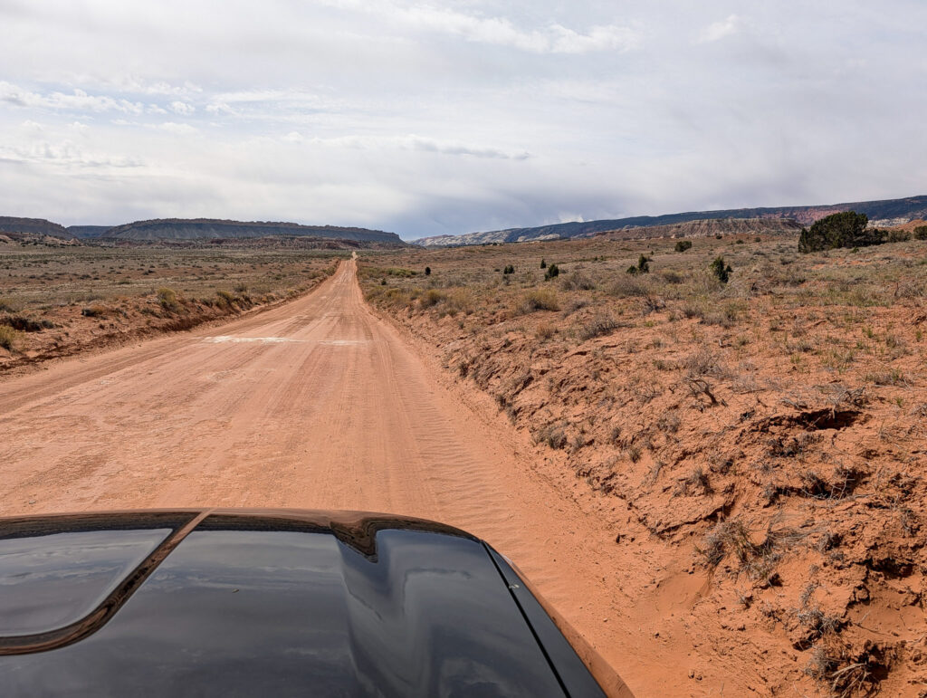 Cottonwood Canyon Road stretches north through red-dirt desert scrubland in Grand Staircase-Escalante, with colorful badlands visible under overcast skies.