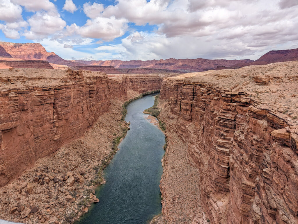 Looking upstream along the Colorado River through Marble Canyon from Navajo Bridge, flanked by vertical red sandstone walls under a partly cloudy sky.