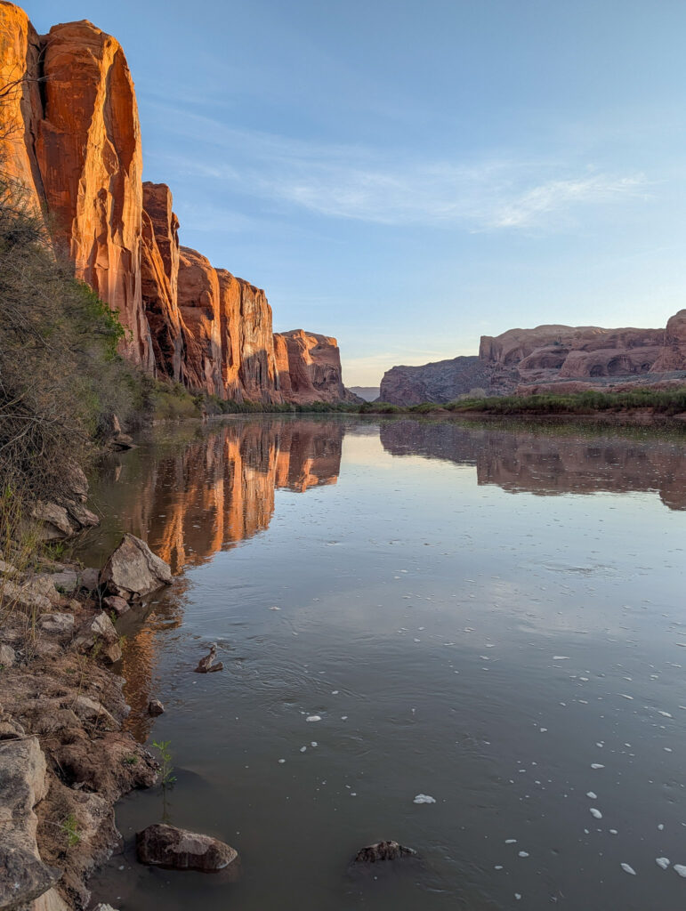 Warm morning light illuminates towering Wingate sandstone cliffs mirrored in the glassy Colorado River near Moab, Utah.