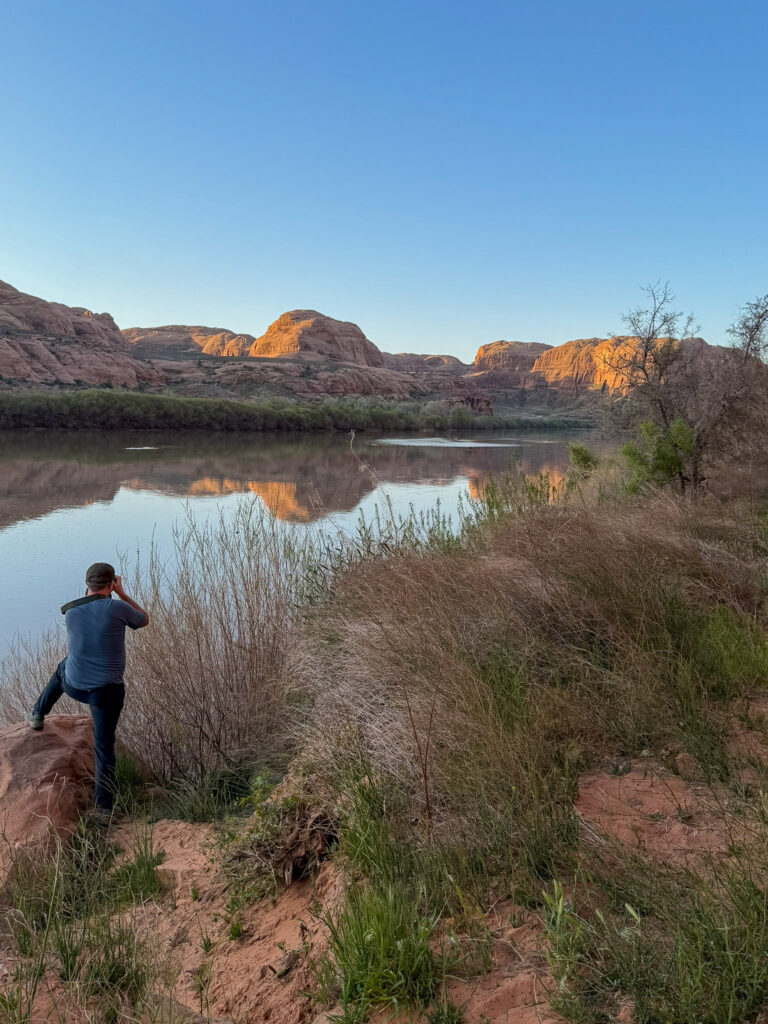 A person stands on a red sandstone bank photographing the Colorado River near Moab as golden-hour light glows on canyon walls reflected in calm water.