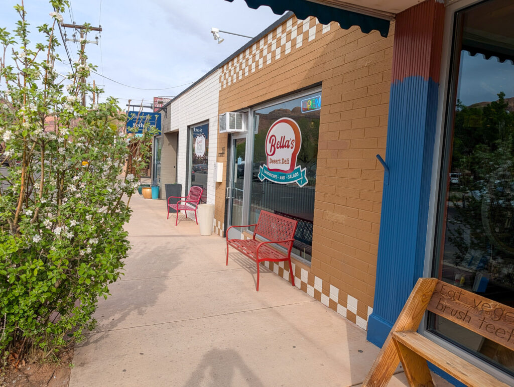 Exterior of Bella's Desert Deli in Moab, with tan brick facade, blue accent pillar, and red bench on the sidewalk.