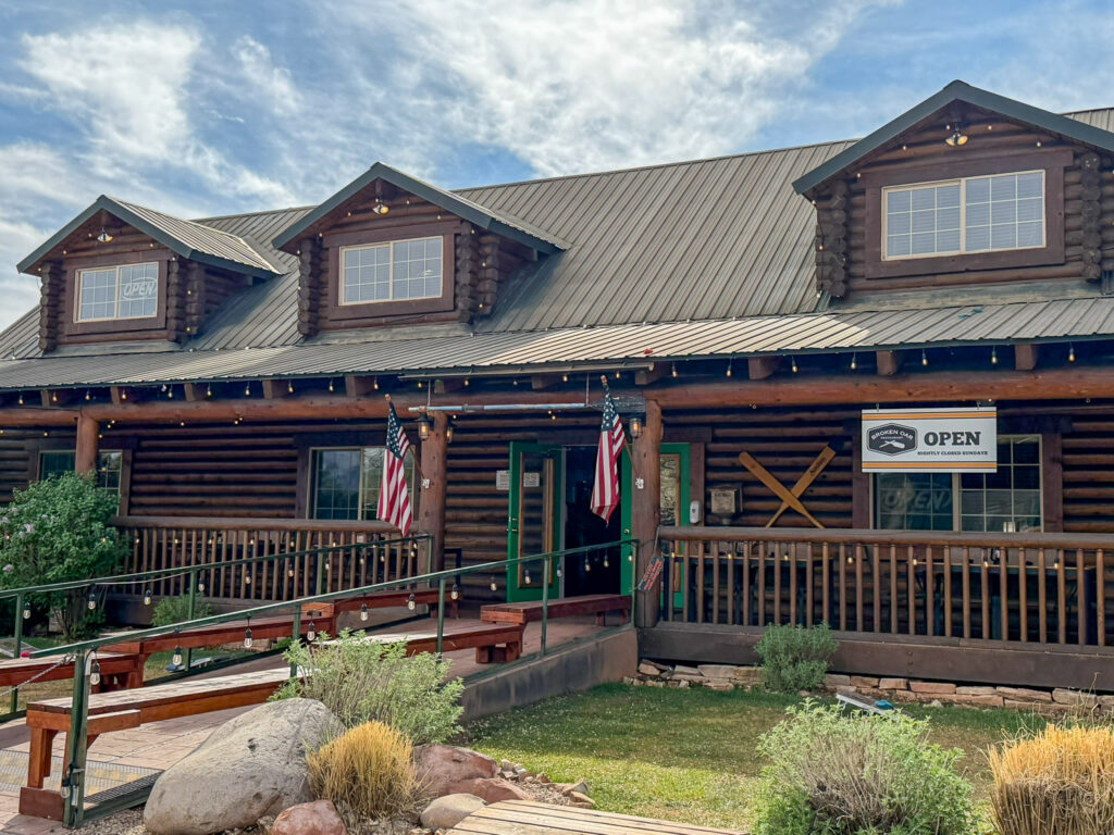 Broken Oar Restaurant's rustic log cabin exterior glows in golden-hour light, with a wraparound porch, string lights, and crossed oar décor.