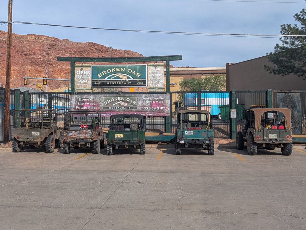 Five muddy Jeeps parked outside the Broken Oar Restaurant in Moab, Utah, with red sandstone cliffs behind.