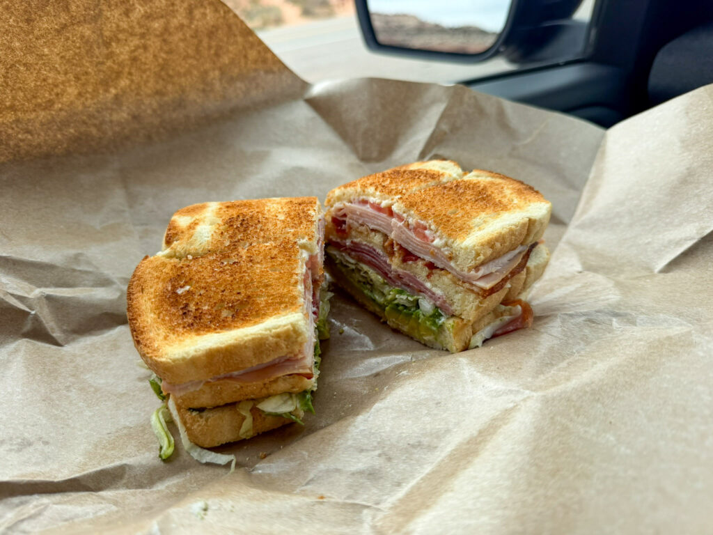 Halved club sandwich on deli paper inside a vehicle, with Moab's red canyon landscape visible through the window.