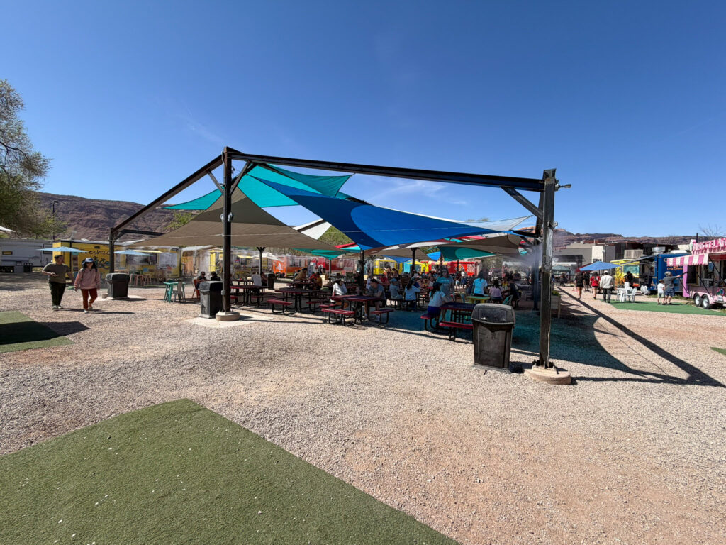 Diners fill picnic tables shaded by colorful sail canopies at the Moab Food Truck Park, with red sandstone cliffs rising behind.