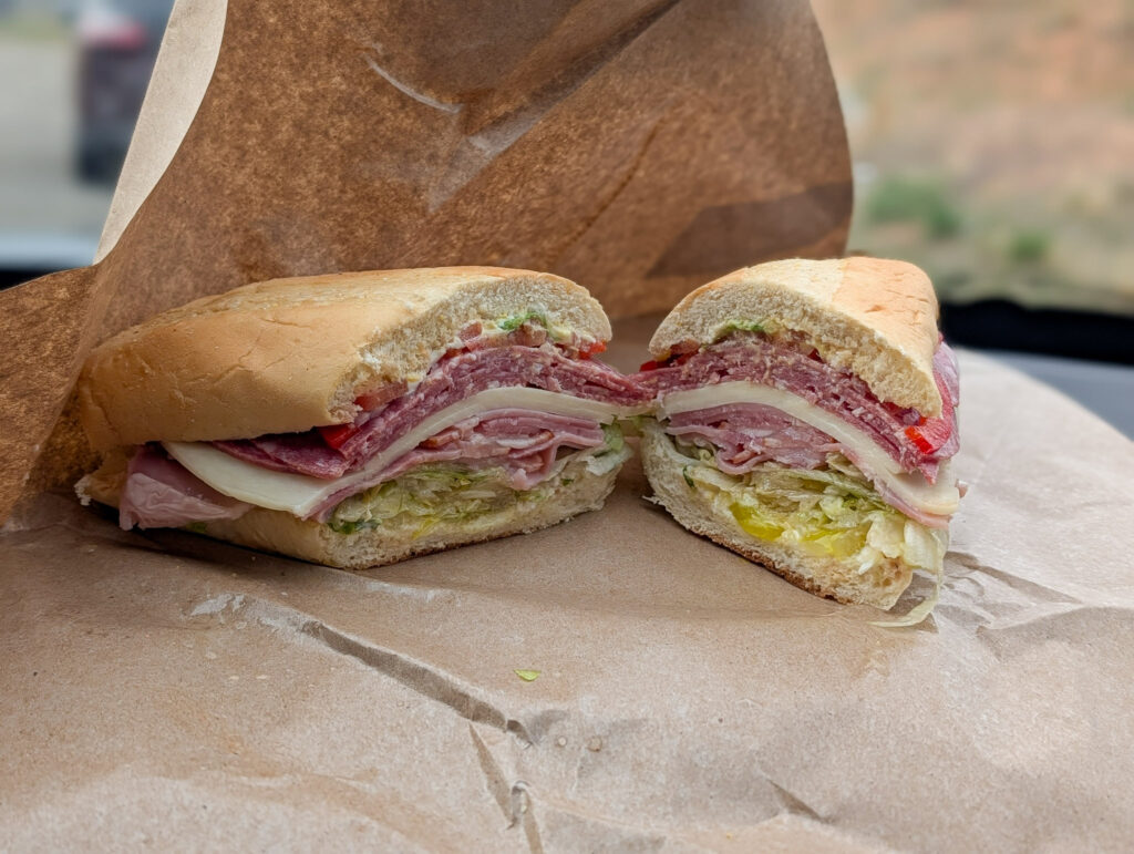 Halved Italian sub sandwich on butcher paper, cross-section showing salami, ham, provolone, lettuce, pepperoncini, and roasted red peppers.