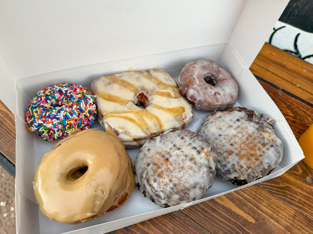 Half-dozen assorted donuts from Moab Donut in an open white box, with orange juice on a wood table nearby.