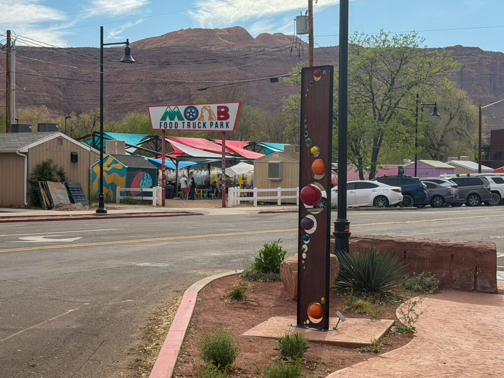 Street-level view of Moab Food Truck Park with colorful canopies, a decorative glass-orb sculpture, and red rock cliffs rising in the background.