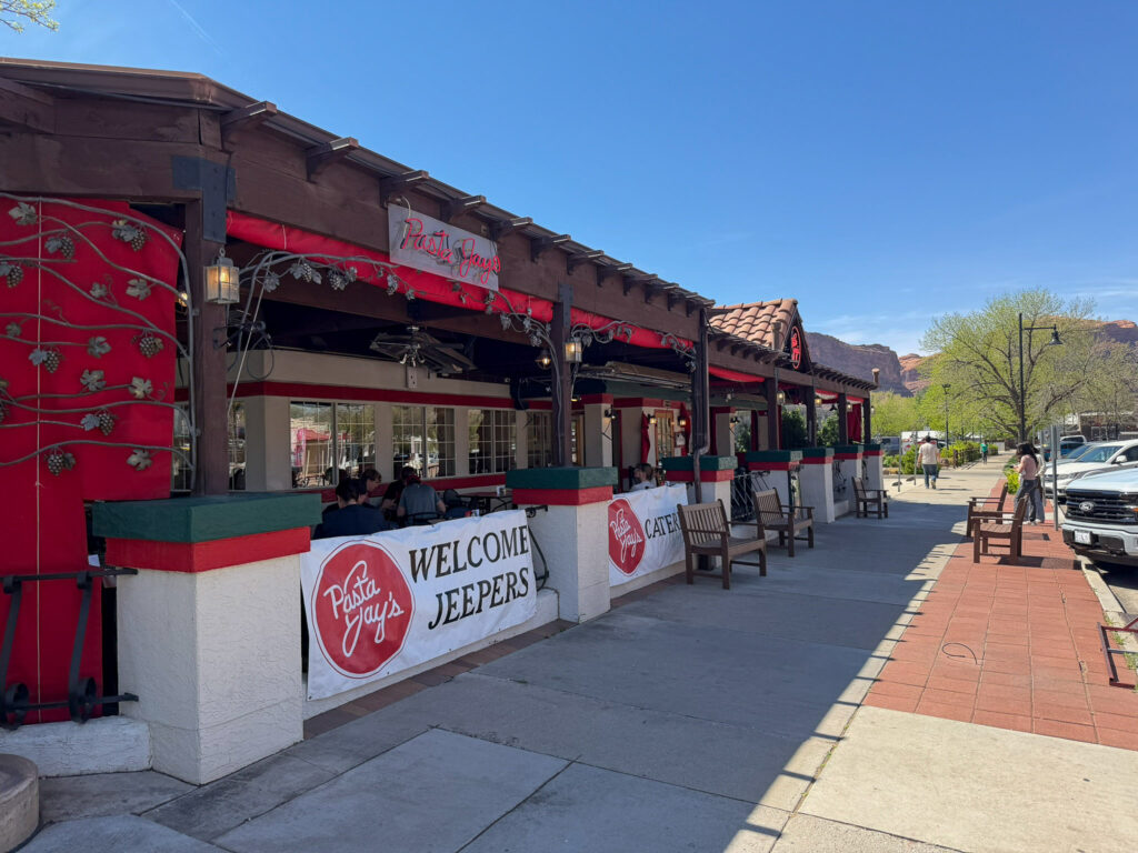 Pasta Jay's restaurant patio in Moab displays a Welcome Jeepers banner beneath red awnings, with canyon cliffs visible beyond.