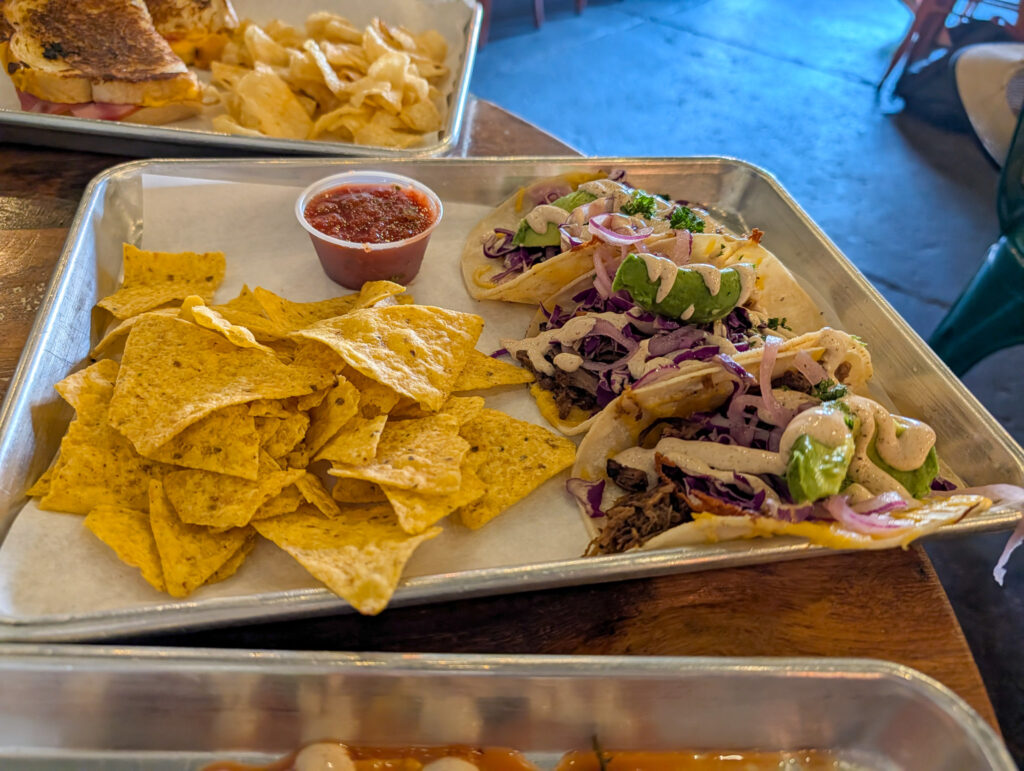 Three flour tortilla tacos topped with cabbage slaw, pickled onions, and avocado, served with chips and salsa on a tray at a casual Moab restaurant.
