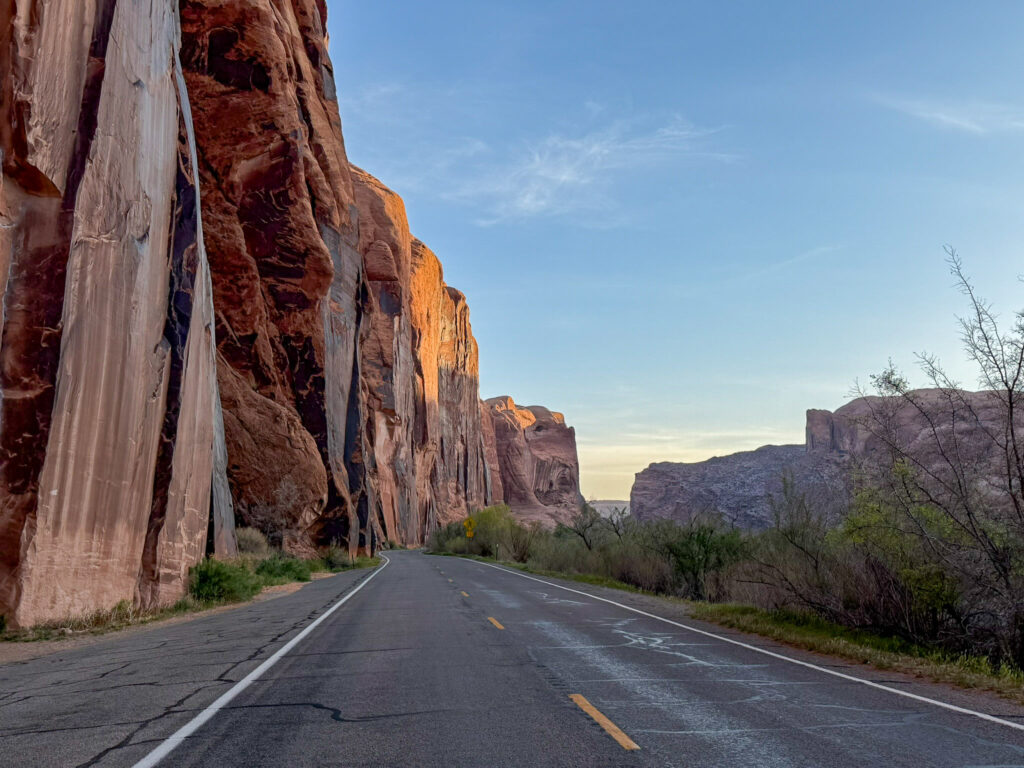 Potash Road curves alongside towering Wingate sandstone cliffs streaked with desert varnish in warm golden-hour light near Moab, Utah.